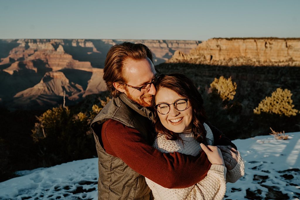 A man hugs a woman from behind at the Grand Canyon and they both smile