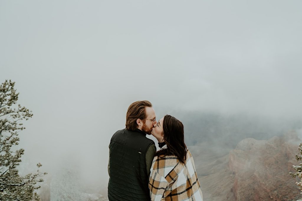Couple kiss in front of fog at the Grand Canyon with their backs turned to the camera