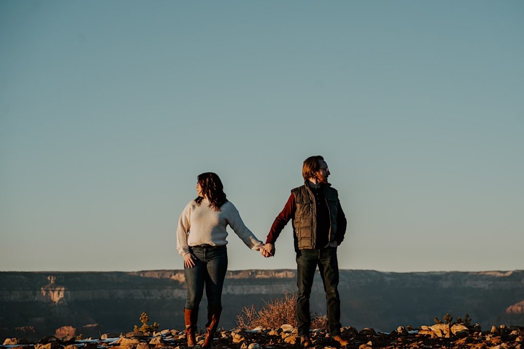 A man and woman stand next to one another and hold hands while looking the opposite direction, the Grand Canyon is behind them