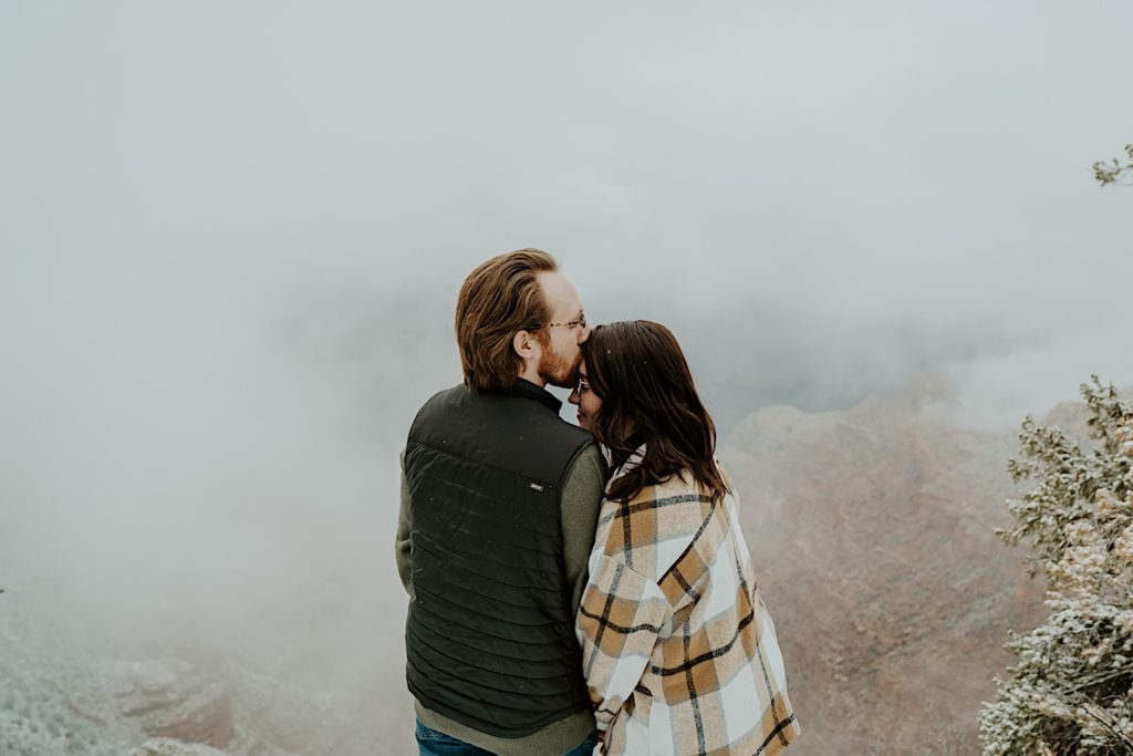 Man kisses woman's forehead in front of fog at the Grand Canyon with their backs turned to the camera
