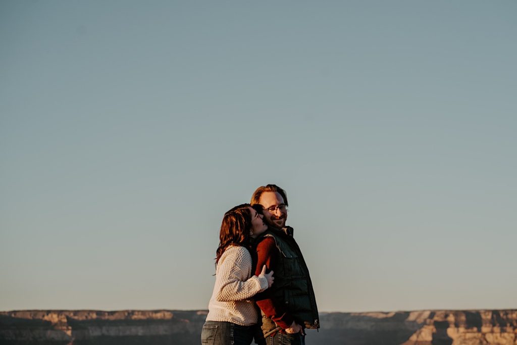 A woman grabs a man by the arm and kisses his cheek from behind as he smiles, the Grand Canyon is in the background