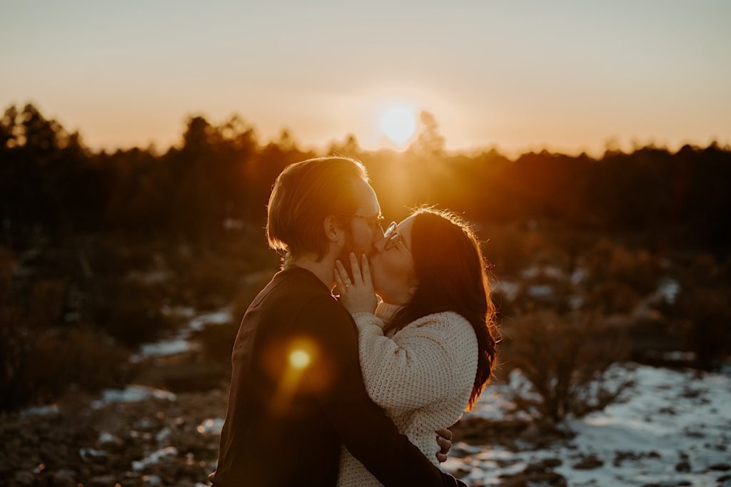 A man and woman kiss one another with a snowy forest and the setting sun behind them
