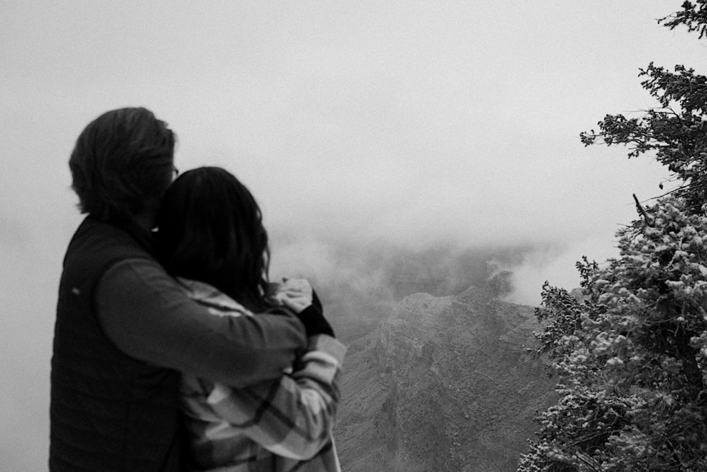 Black and white photo of a man holding a woman from behind while they look out over the foggy Grand Canyon together