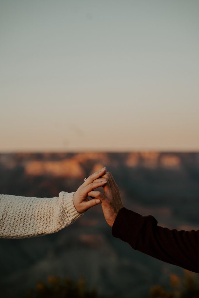 Two hands extend and touch one another in front of the Grand Canyon, one of the hands has an engagement ring on it
