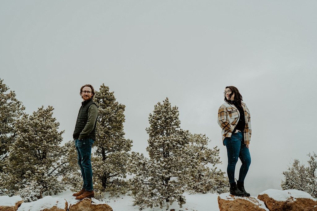 A man and woman stand on separate rocks with their backs turned to one another, they are turning to look at one another while the snow falls around them at the Grand Canyon