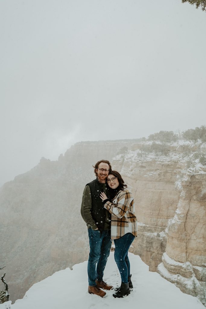 Couple stand next to one another and smile at the camera as snow falls around them with the Grand Canyon in the background