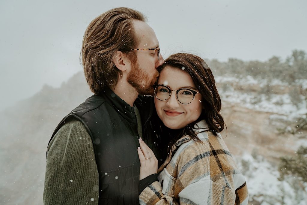 Couple stand next to one another in front of a snow covered Grand Canyon, the man kisses the woman on the temple while she smiles at the camera and shows off her engagement ring by putting her hand on his chest