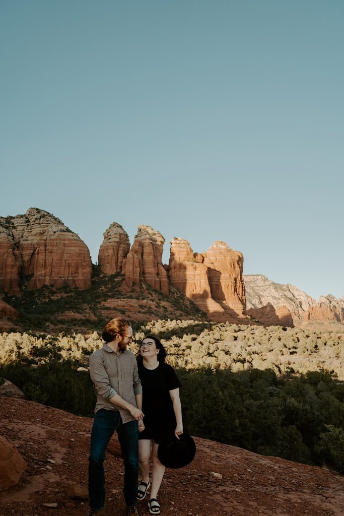 A couple walk towards the camera holding hands and smiling at one another with Sedona red rocks in the background