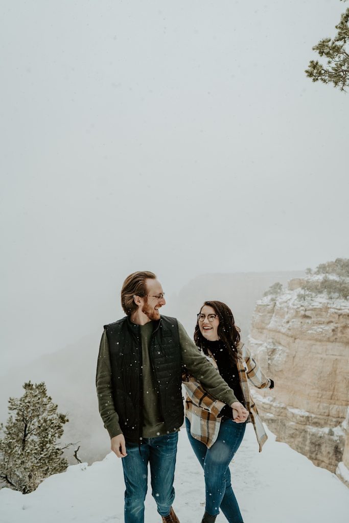 Couple hold hands and look at one another while smiling and walking towards the camera, there is snow falling around them and the Grand Canyon is behind them