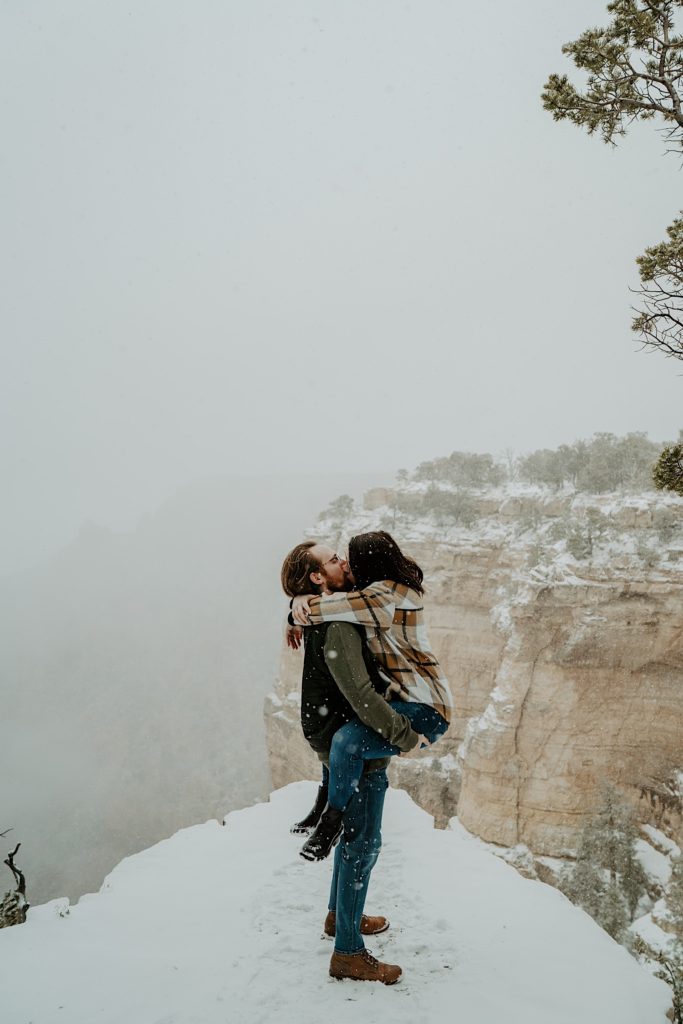 Couple standing on a cliff kiss as the man lifts the woman by the waist, there is snow falling around them and the Grand Canyon is in the background