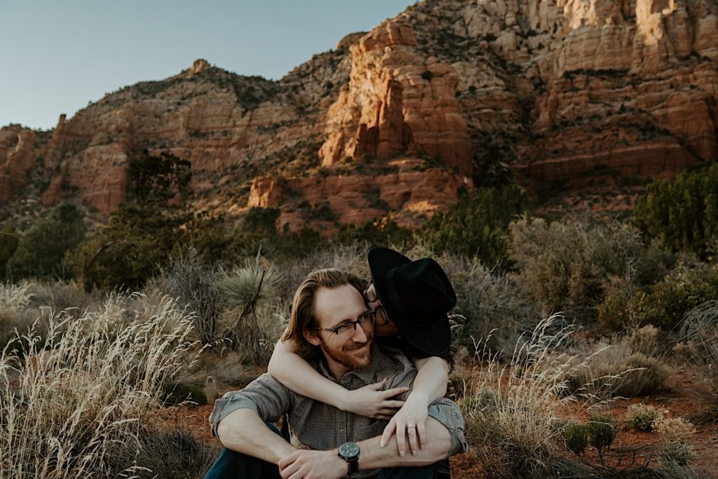 A man sits in the desert in front of the Sedona red rocks and smiles as his fiancée embraces him from behind and kisses his cheek