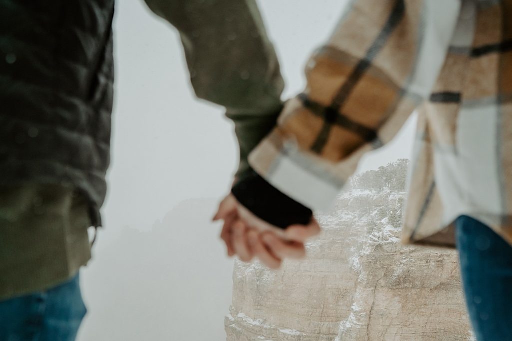 Close up of a couple's hands interlocked, their hands are blurry while the snow covered Grand Canyon is in focus in the background
