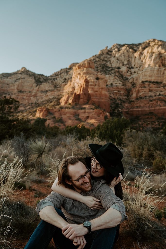 A man sits in the desert in front of the Sedona red rocks and smiles as his fiancée embraces him from behind