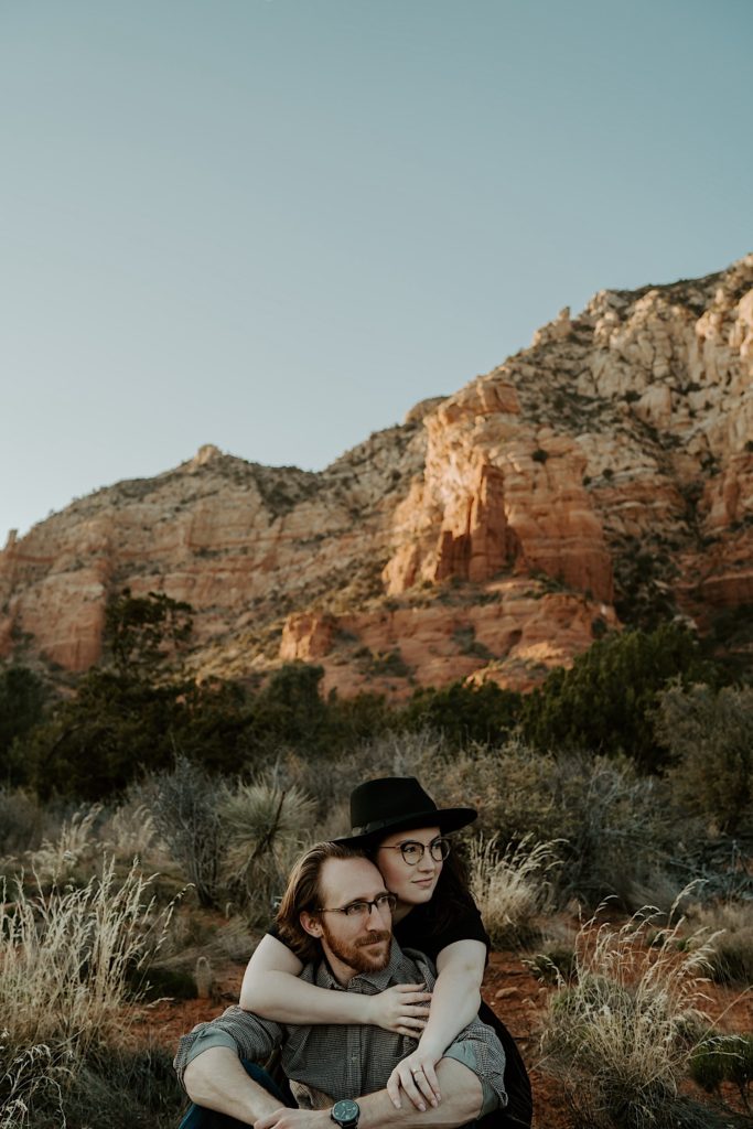 A couple sit in the desert with the Sedona red rocks behind them, the woman is behind the man with her arms around him as they both look to the right