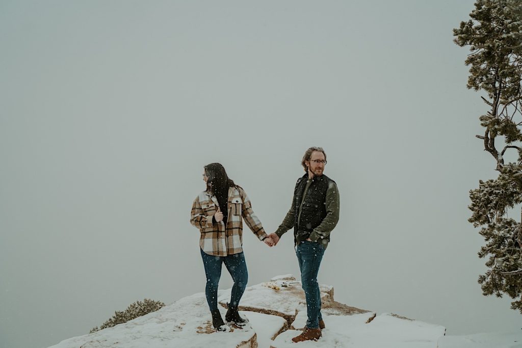 A couple stand on a snowy rock holding hands and looking opposite directions from one another with dense fog/snow behind them