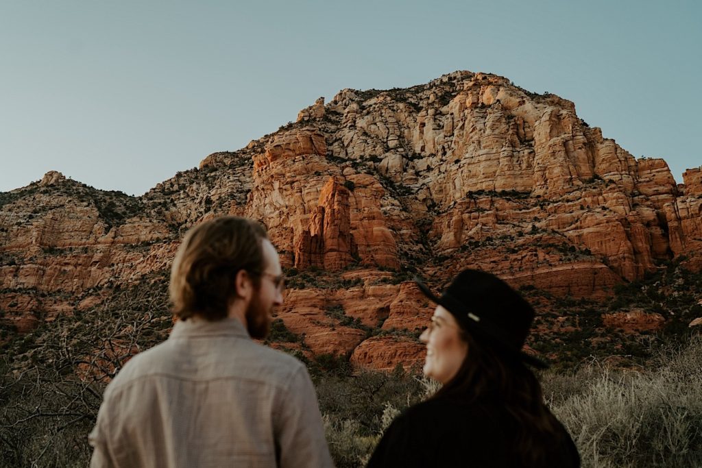 A couple stand with their backs turned to the camera looking at one another and smiling, the couple is blurry while the Sedona red rocks in the background are in focus
