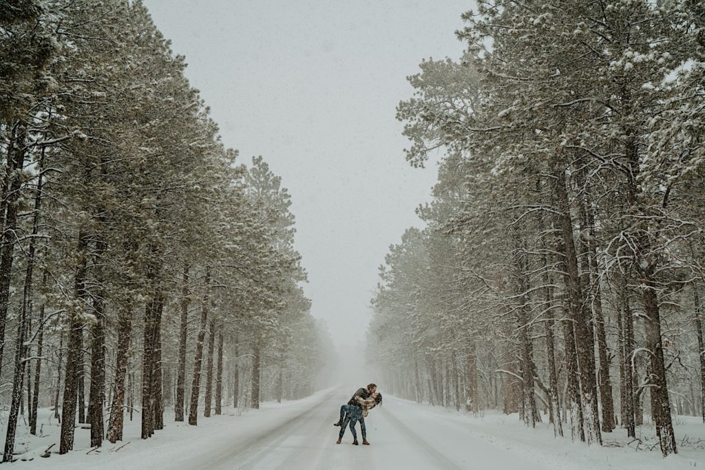 A couple kiss in the middle of a road, the road is in the middle of a forest with tall trees on either side of it, snow is falling and covering the road and trees
