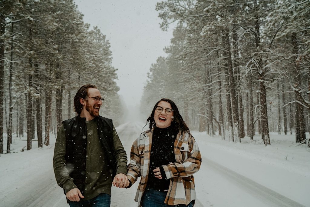 A couple walk towards the camera while laughing and holding hands in the middle of a road, the road is in the middle of a forest with tall trees on either side of it, snow is falling and covering the road and trees