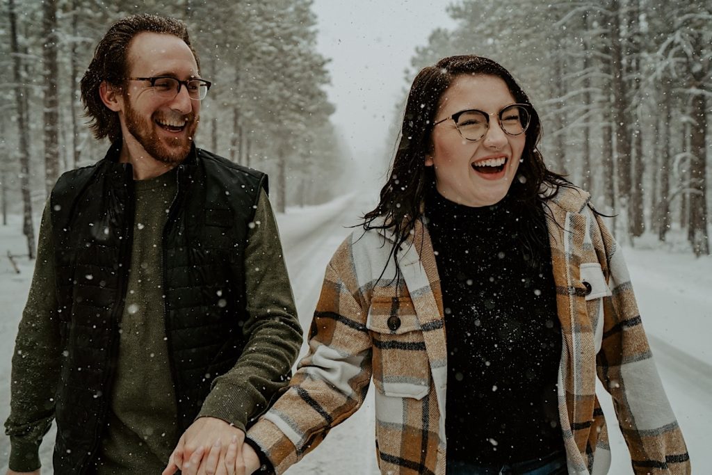 A couple walk towards the camera while laughing and holding hands in the middle of a road, the road is in the middle of a forest with tall trees on either side of it, snow is falling and covering the road and trees