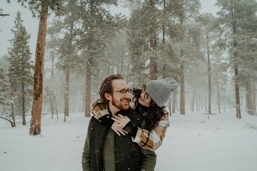 A woman hugs a man from behind in a snowy forest, they smile and look at one another