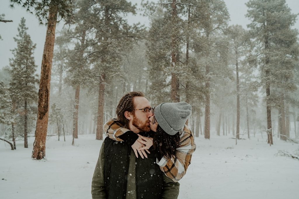 A woman hugs a man from behind and they kiss one another in a snowy forest