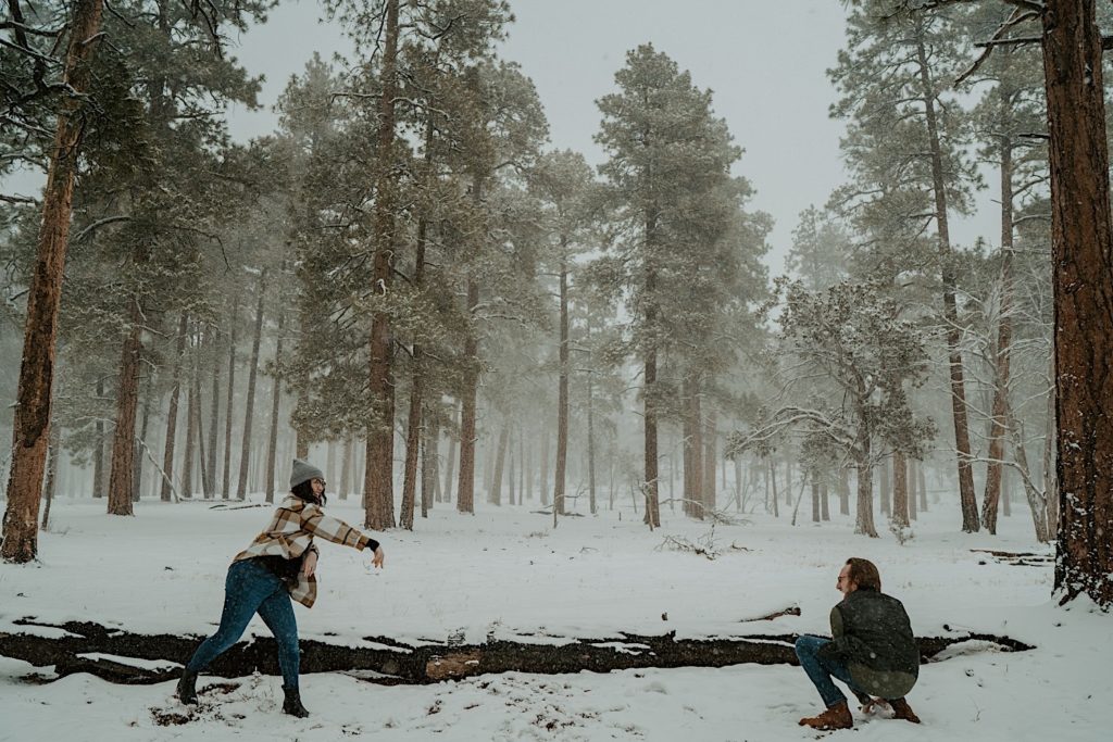 A man and a woman have a snowball fight in a snow covered forest, the woman is throwing a snow ball while the man is crouched making his own snow ball