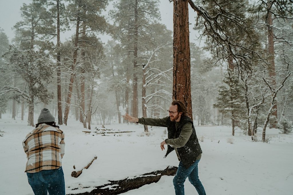 A man and a woman have a snowball fight in a snow covered forest, the man is throwing a snowball at the woman
