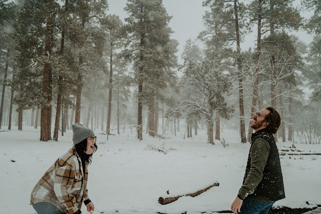 A man and a woman stand facing one another laughing extremely hard in the middle of a snowy forest