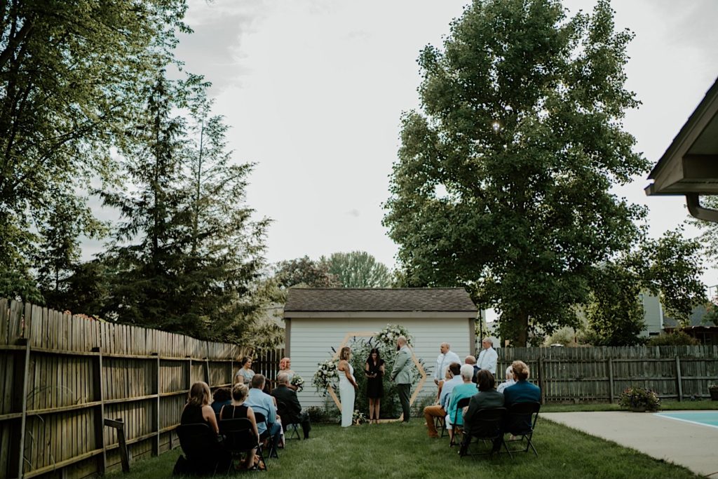 Bride and groom standing next to their priest while their guests are seated during their backyard wedding ceremony