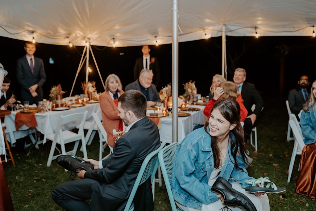 Bride and groom are seated back to back while holding one of each of their shoes and laughing under a tent with their guests during their wedding reception
