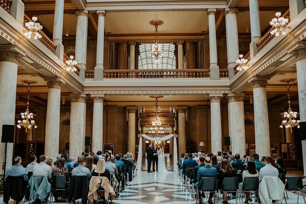 Bride and groom hold hands at their wedding ceremony at the Indiana State House while their guests are seated watching