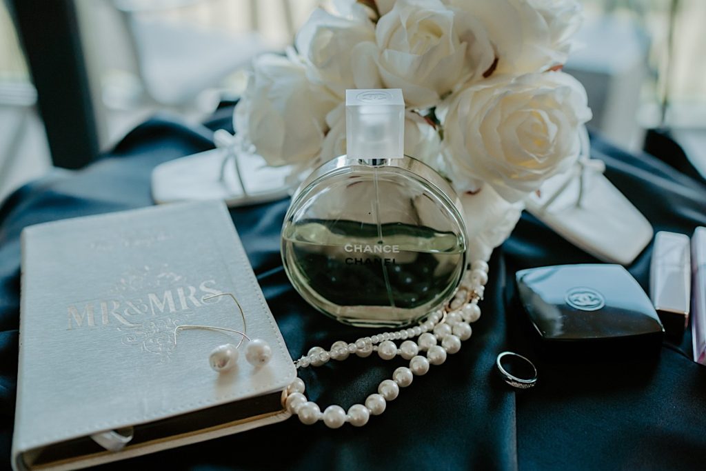 Close up photograph of perfume, a necklace and earrings, a wedding ring, flowers and a book with "MR&MRS" engraved on it.