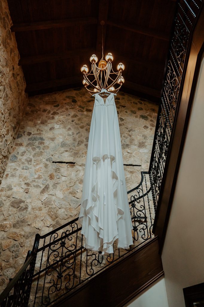 Photo looking up at a wedding dress handing from a chandelier with a staircase and stone walls around it.