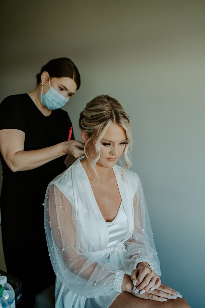 A bride sits in a white robe as a hair stylist stands behind her working on her hair.