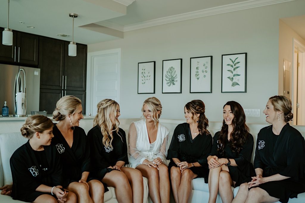 A bride sits on a couch with her bridesmaids before they get ready for a wedding, the bridesmaids are wearing black robes while the bride wears a white robe