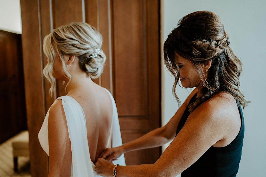 A bride stands facing away from the camera as a woman helps button up the back of her wedding dress.