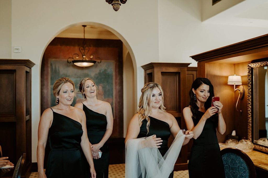 Four bridesmaids stand and smile while waiting for the bride to enter wearing her wedding dress, one bridesmaid is holding her phone to take a photo while another holds the bride's veil.