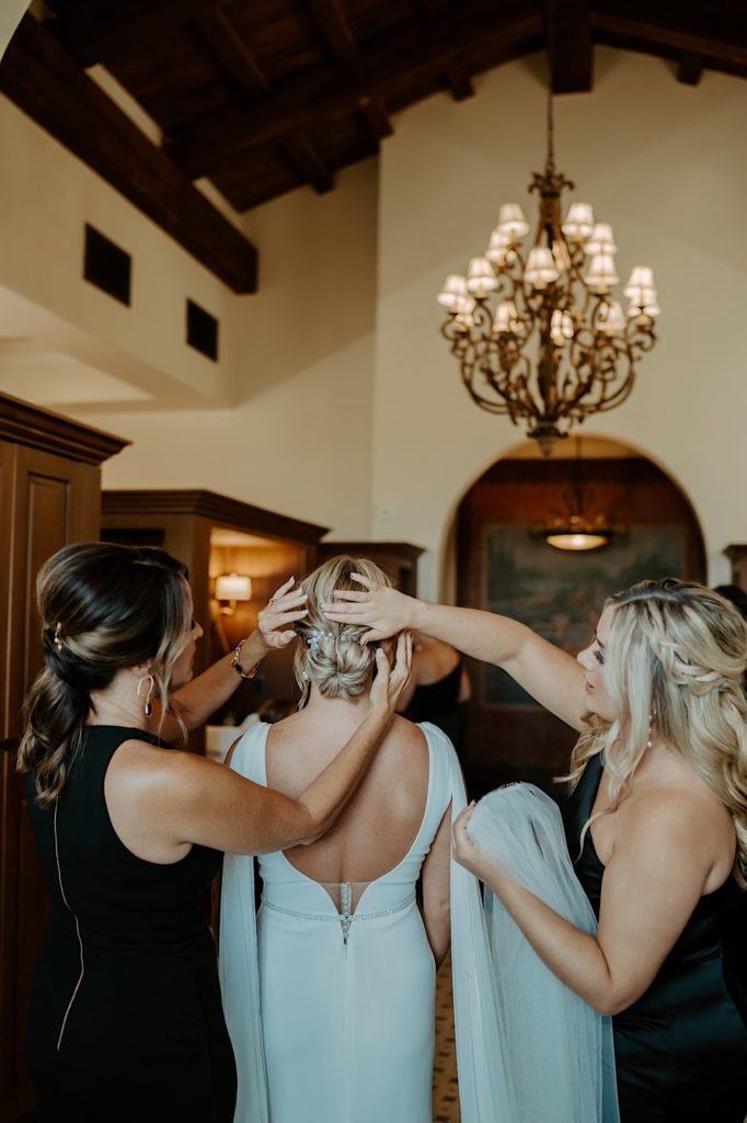 A bride stands between two bridesmaids with her back to the camera as the bridesmaids adjust her hair.