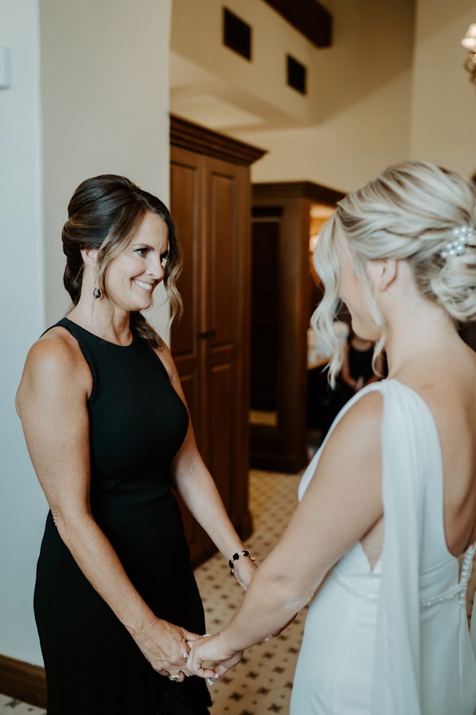 A bride stands facing one of her bridesmaids who is smiling back at her as they hold each others hands.