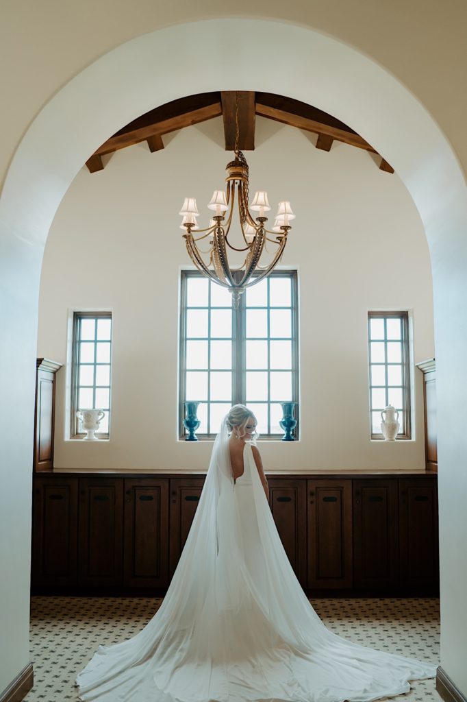 A bride in her wedding dress stands underneath a chandelier in an archway with her back facing the camera 