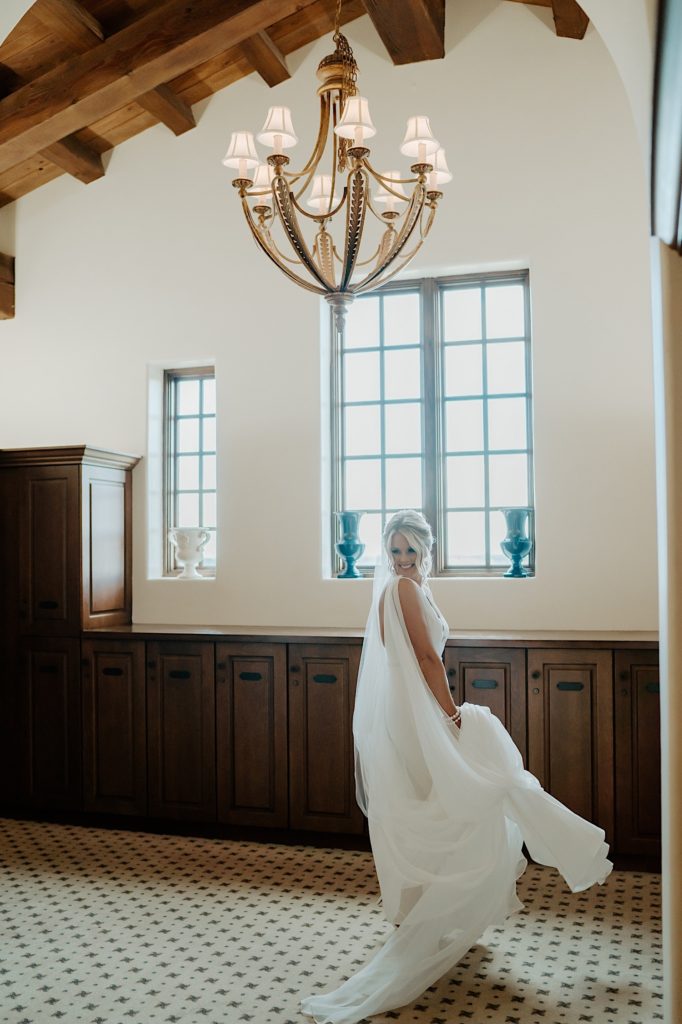 A bride in her wedding dress turns towards the camera and smiles underneath a large chandelier.