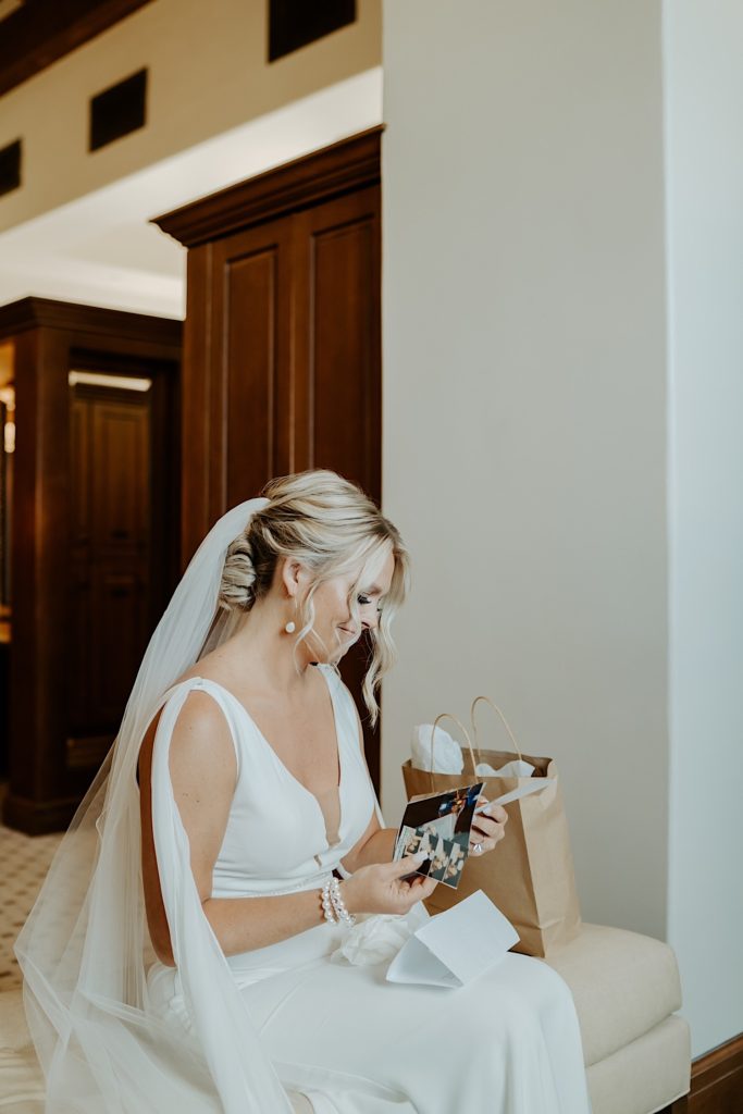 A bride in her wedding dress sits and smiles as she looks through photographs from a gift bag from her soon to be spouse.