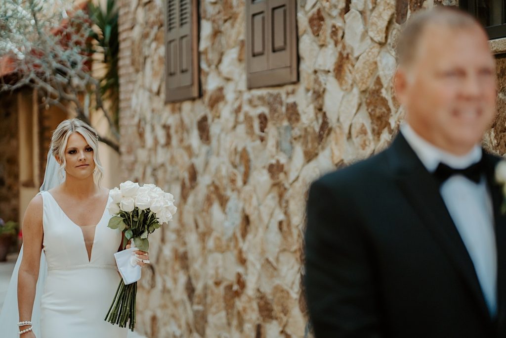 A bride holding a flower bouquet walks towards her father, who has his back turned to her and is out of focus in the photo, to surprise him for a first look.