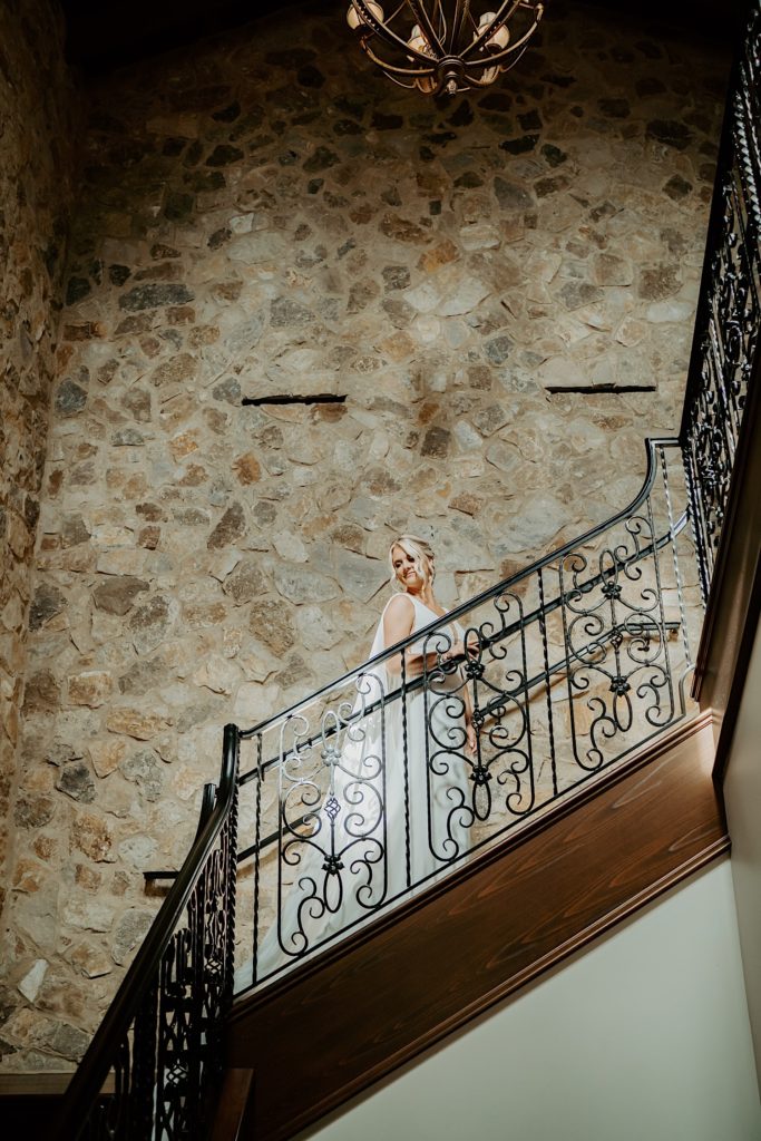 A bride in her wedding dress stands on a staircase with a stone wall behind her and a chandelier above her