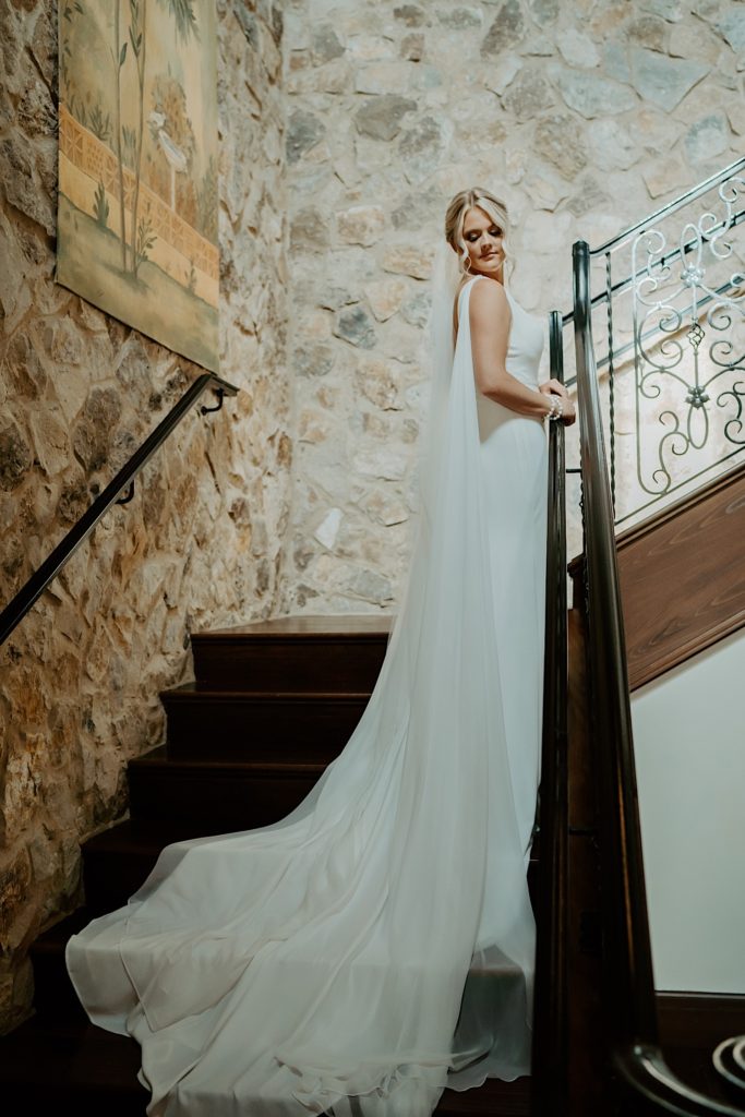 A bride in her wedding dress stands on a staircase with a stone wall behind her.