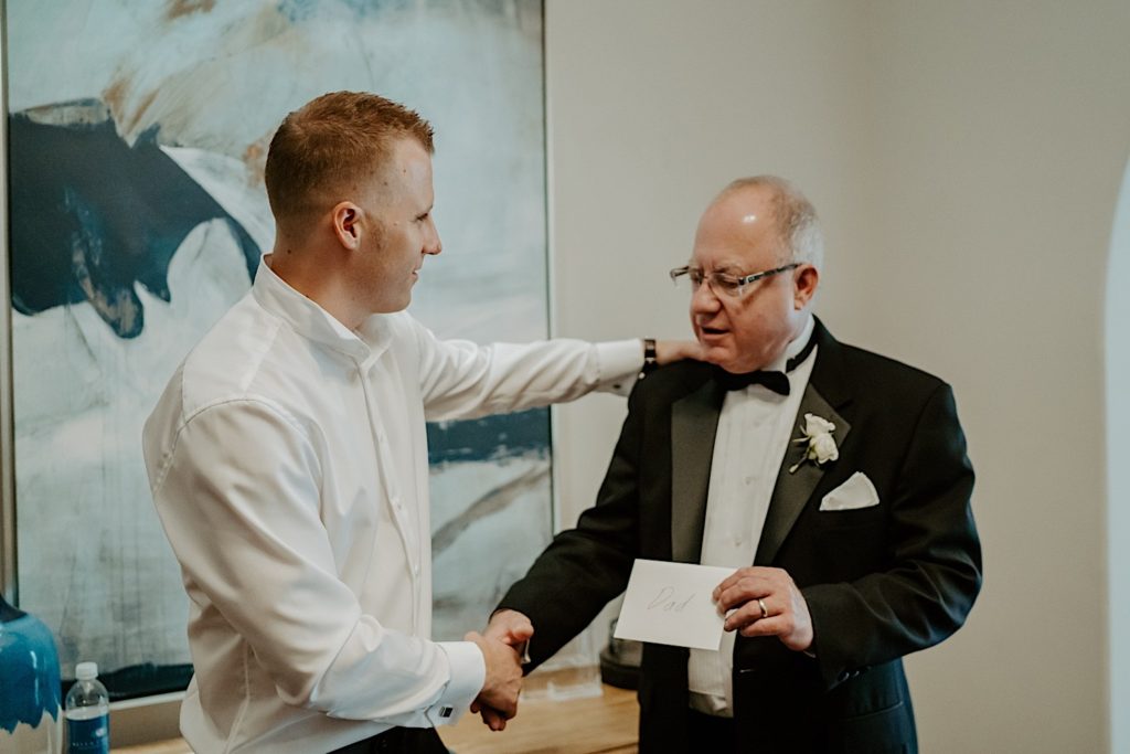 A groom stands and shakes his fathers hand before his wedding day, the father is holding an envelope that reads "Dad"