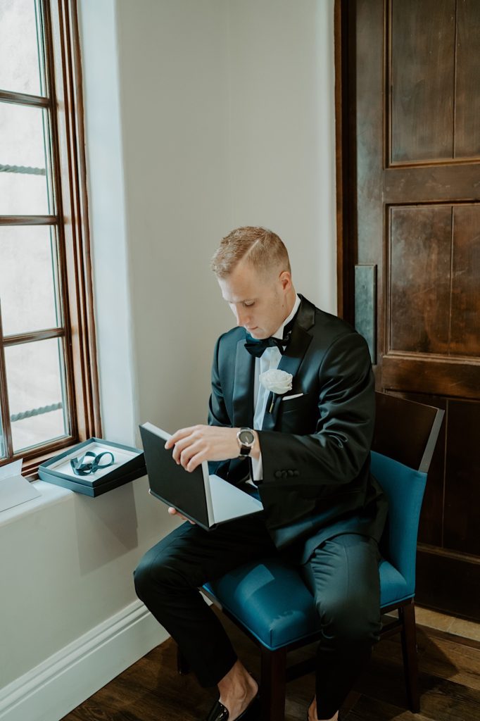 A groom dressed for his wedding sits and opens a book that is a gift from the bride