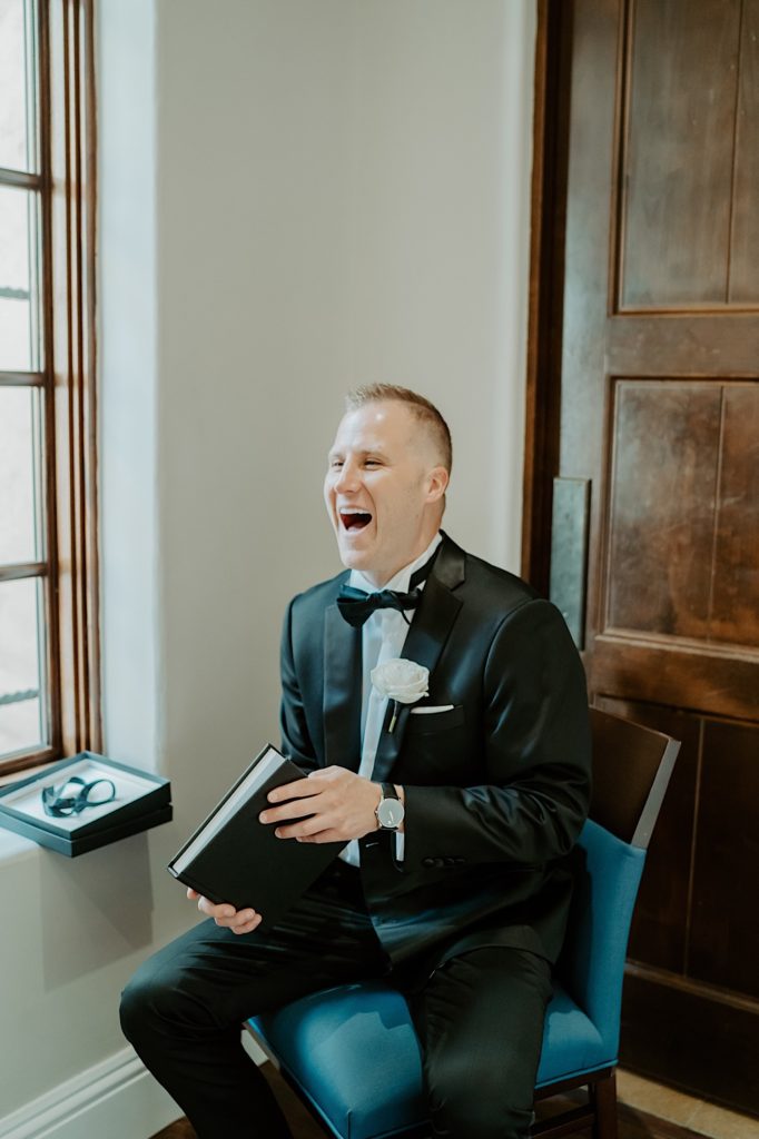A groom dressed for his wedding sits and laughs as he opens a book from the bride.