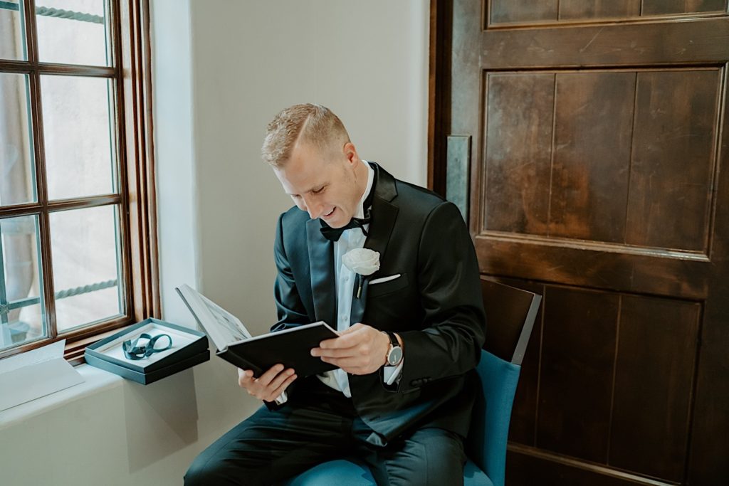 A groom dressed for his wedding sits and smiles as he looks through a book that was a gift from the bride.