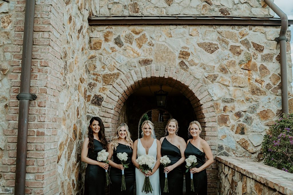 A bride stands with her 4 bridesmaids, 2 on either side of her, all dressed for the wedding and holding bouquets of white roses underneath a stone archway at their intimate Florida wedding venue